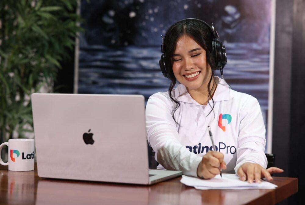 A smiling virtual assistant wearing headphones, writing notes and working on a laptop, showing what do real estate virtual assistants.