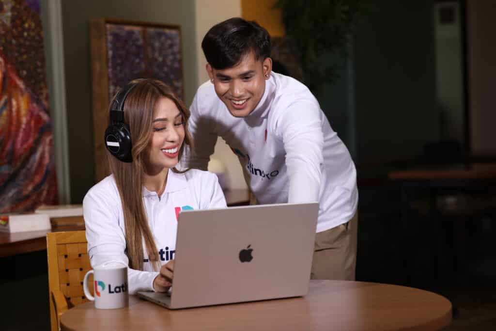 A woman with headphones works on a laptop while a male colleague offers guidance. Both wear LatinoPro shirts and appear focused and collaborative in a modern workspace, representing digital teamwork.