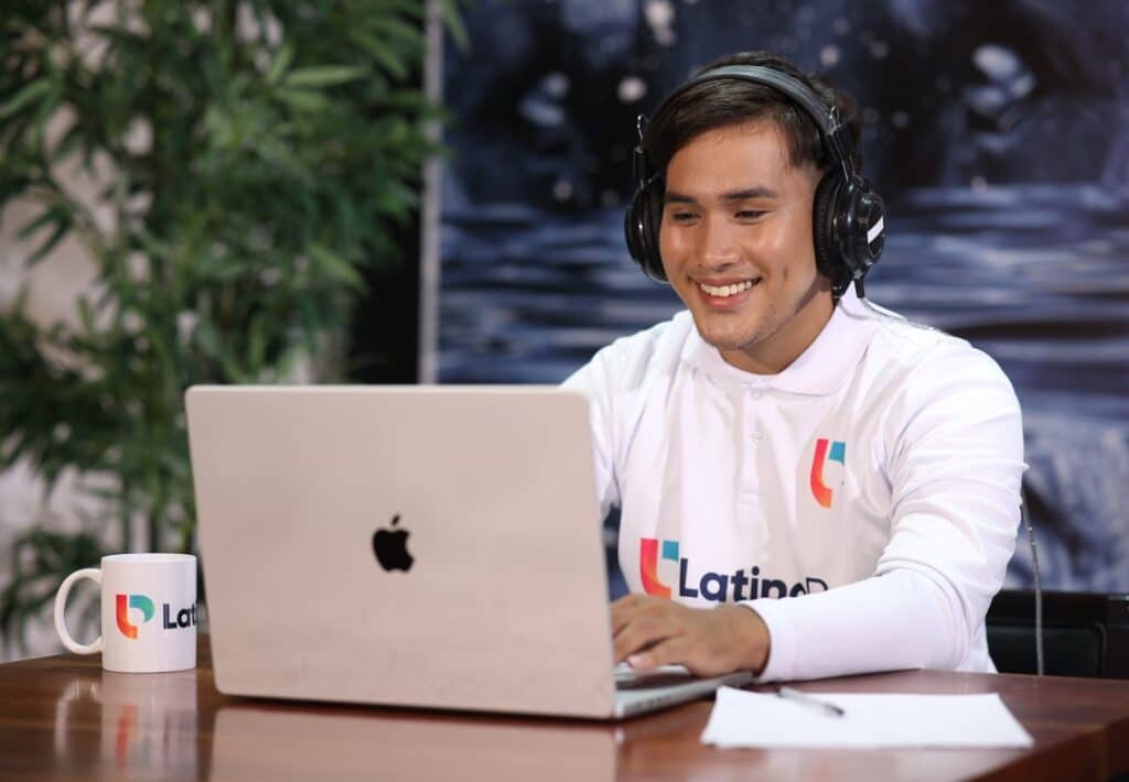 Happy Latino Pro team member with headphones typing on a laptop, with a branded mug beside him