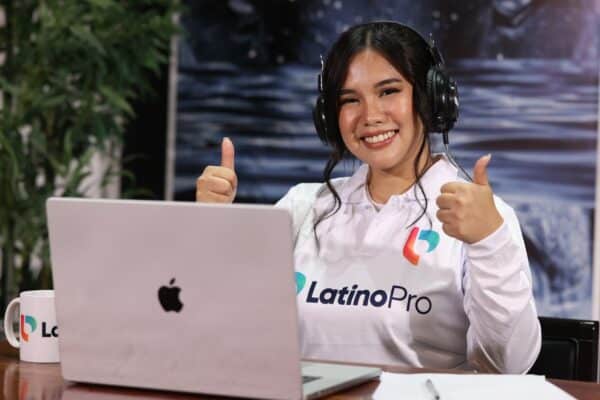 Happy woman with headphones and LatinoPro shirt, giving thumbs up, at a laptop, embodying efficient executive assistant duties
