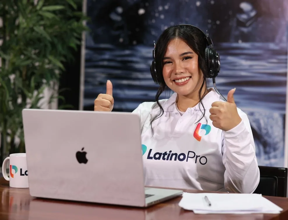 Happy woman with headphones and LatinoPro shirt, giving thumbs up, at a laptop, embodying efficient executive assistant duties