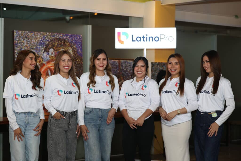 Six women in branded LatinoPro shirts stand together, smiling confidently in a modern office space with artwork and the company logo in the background—conveying unity, professionalism, and team spirit.