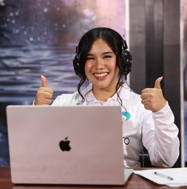 Woman smiling with headphones on, giving two thumbs up while sitting at a laptop