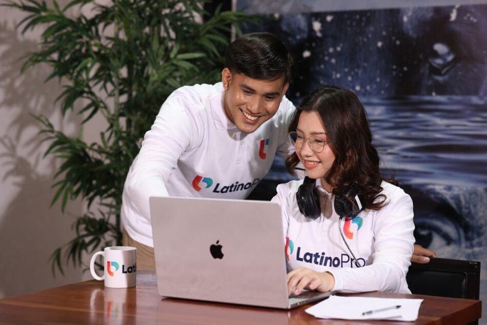 A man and woman smiling while working together on a laptop. They are both wearing white long-sleeve polo shirts.