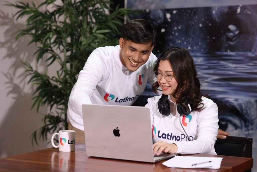 Two smiling young professionals in branded shirts collaborate on a laptop. A mug and papers are on the desk. This could represent teamwork and studying for a legal assistant certification.